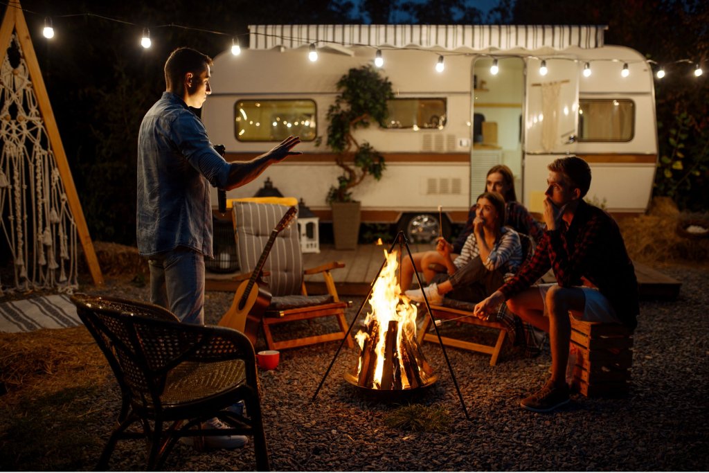 A group of friends making s'mores around a fire and hanging out outside of a lit-up RV at night