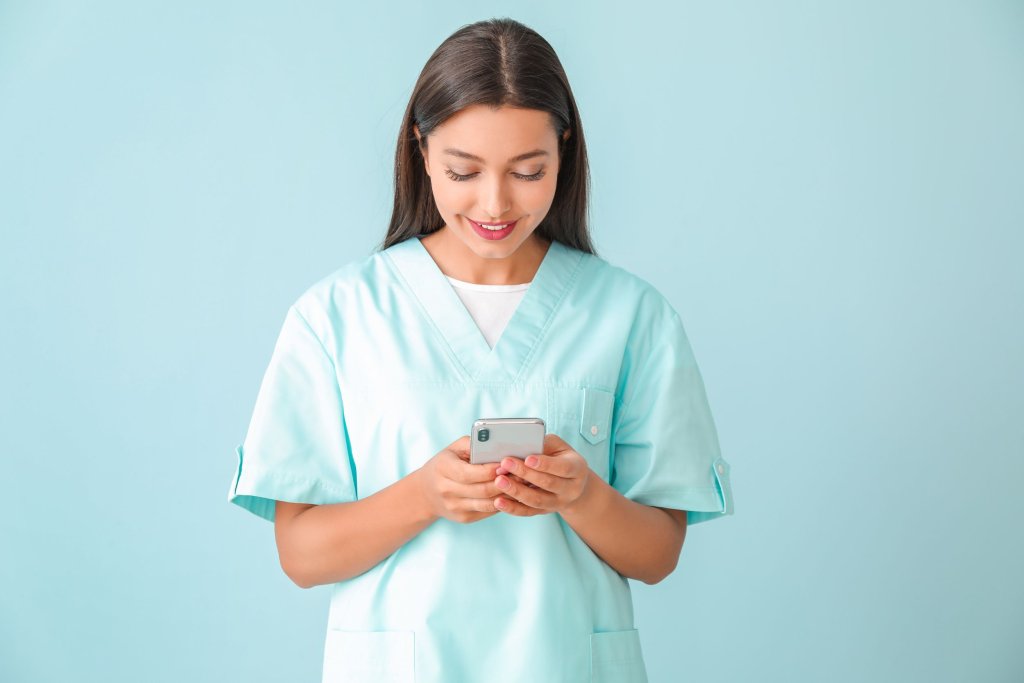 A nurse in scrubs in front of a blue background looking at her phone