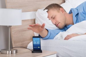 man lying in bed sleeping, reaching for his alarm on his phone.